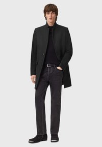 Musta takki, jossa on korkea kaulus ja nappidetali, yhdistetty tummiin farkkuihin ja nilkkabootseihin. Sileä pinta, moderni muotoilu, vähäiset yksityiskohdat.