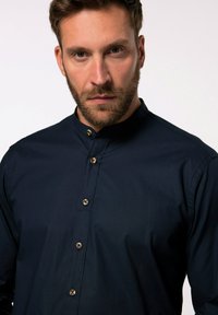 Camicia blu navy con colletto rialzato, caratterizzata da bottoni marroni e un tessuto liscio e testurizzato. Il design è minimalista e aderente.