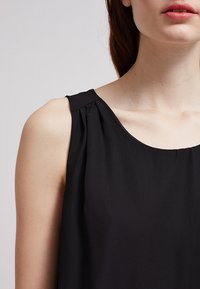 Černý bezrukávový top vyrobený z lehkého, texturovaného materiálu. Má kulatý výstřih a na rameni nařasený detail.