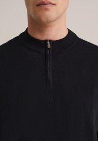 Pull-over en maille foncée avec un col montant et une fermeture éclair frontale à moitié, présentant une texture lisse et un design minimaliste.