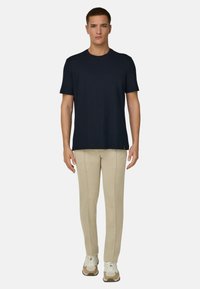 Homme debout, de face, portant un t-shirt noir uni à manches courtes, un pantalon beige avec des coutures frontales, et des baskets beige et blanches.