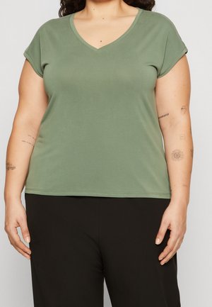 Osoba o zaokrąglonych kształtach ubrana w oliwkowo-zielony T-shirt z krótkim rękawem i dekoltem w serek oraz czarne spodnie, pokazująca tatuaże na obu przedramionach.