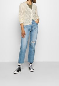 Kremowy sweter z dzianiny z dekoltem w serek, zapinany na guziki, z fakturowanym wzorem, zestawiony z jasnoniebieskimi, przetartymi jeansami o prostych nogawkach oraz czarnymi trampkami za kostkę.