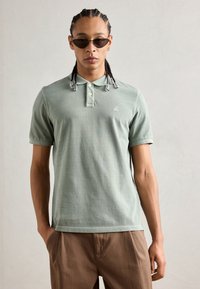 Polo-skjorte i myk grønn bomull, med to-knapp placket, korte ermer og en liten brodert logo på brystet. Brukt med beige bukser.