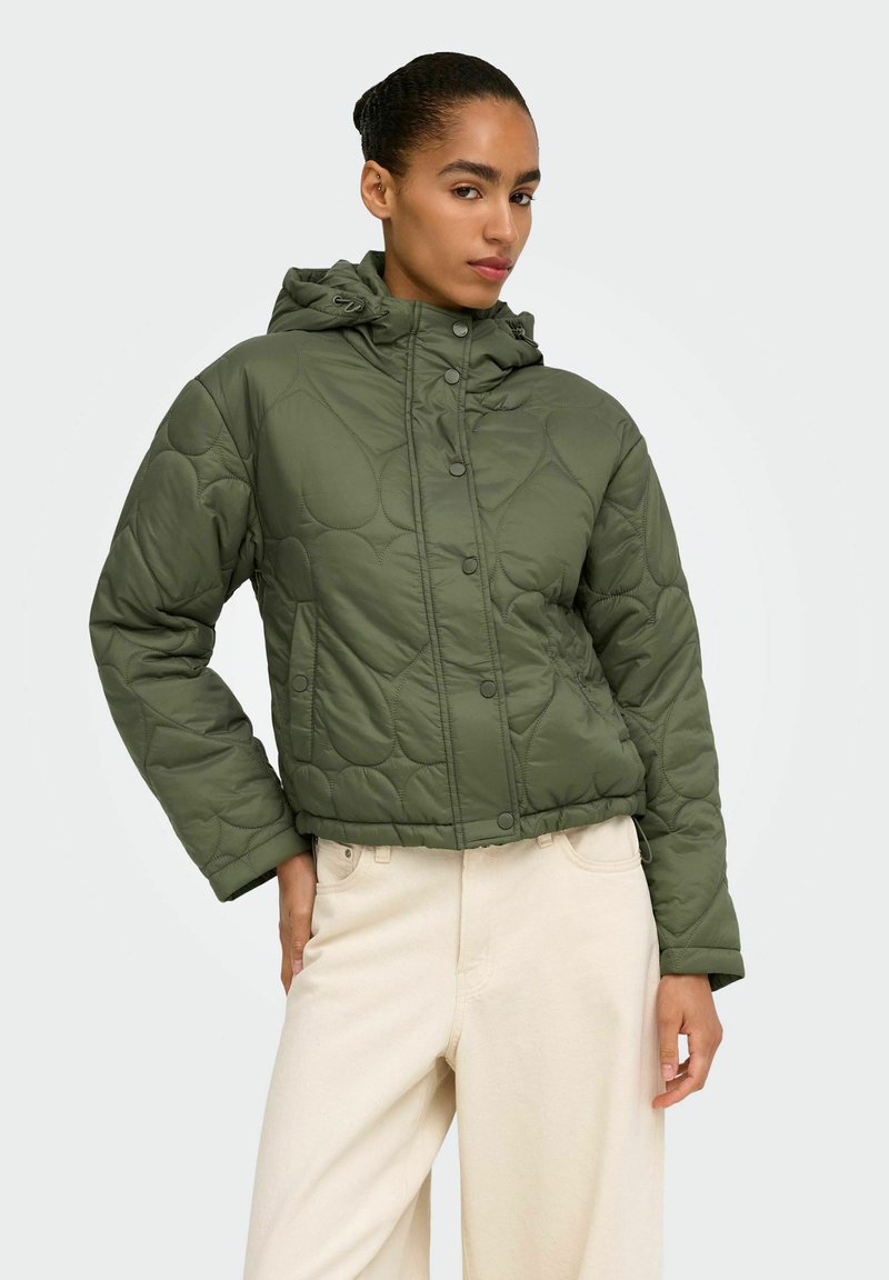 Femme portant une veste matelassée à capuche vert olive et un pantalon beige clair, debout avec une main sur la hanche devant un fond uni.
