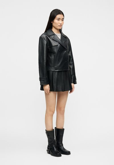 Γυναίκα με μαύρο δερμάτινο σακάκι διπλής σειράς κουμπιών, πλισέ δερμάτινη μίνι φούστα, μαύρες μπότες μέχρι τη μέση της γάμπας, στέκεται μπροστά σε λευκό φόντο.