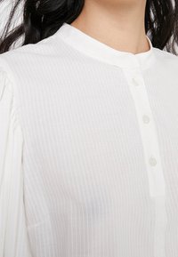 Camicia bianca a righe con colletto alla cinese, dotata di chiusura frontale a bottoni e texture morbida e leggera. Maniche lunghe, leggermente arricciate.