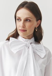 Blusa blanca con un gran lazo en el cuello, hecha de tela suave. Presenta un corte relajado y mangas largas. Pendientes de aro de oro visibles.