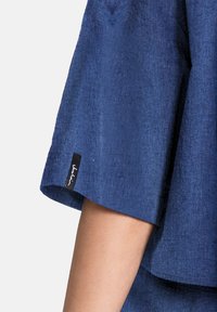 Blå denimskjorta med korta, utsvängda ärmar. Har en subtil struktur och en svart etikett på fållen med vit sömnad.