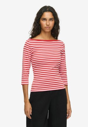 Femme aux cheveux foncés ondulés portant un haut rayé horizontal rouge et blanc avec un col bateau et un pantalon noir sur fond blanc uni.