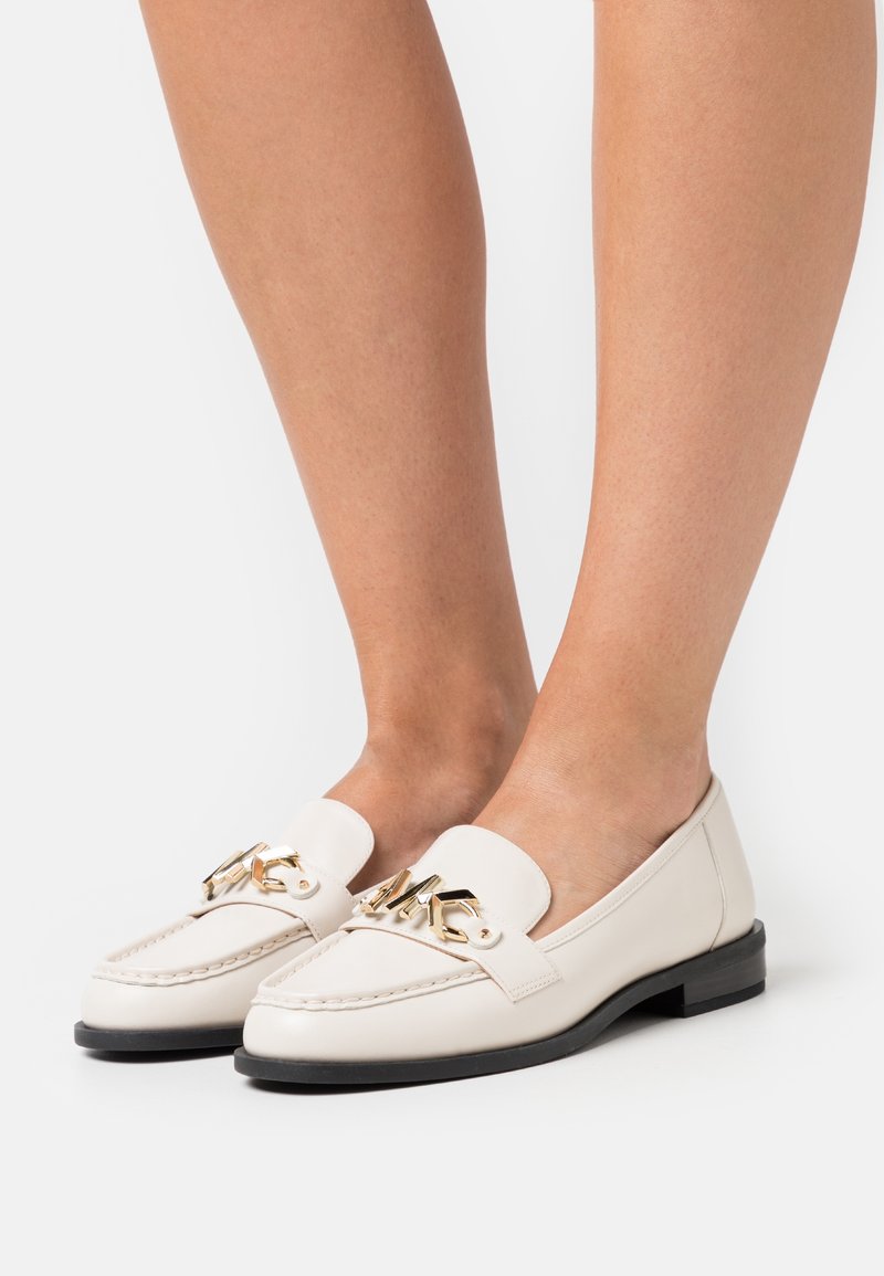 Pieds portant des mocassins en cuir blanc avec des accents dorés du logo "MK" et des semelles noires sur fond blanc.