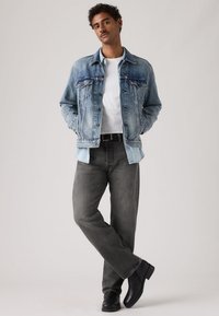 Νεαρός άνδρας στέκεται με τα χέρια στις τσέπες, φοράει μπλε τζιν μπουφάν, άσπρο πουκάμισο, γκρι τζιν και μαύρα μποτάκια σε απλό φόντο.