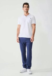 Camisa polo blanca con un pequeño emblema en el pecho, combinada con pantalones azul marino y zapatillas blancas, presentada en una posición de pie.