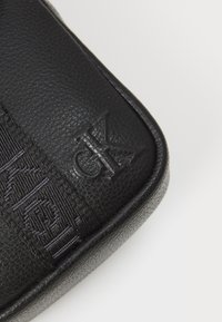 Schwarze Lederhandtasche mit strukturiertem Oberflächen, geprägt mit Logo und einem gewebten Tragegurt. Abgerundete Kanten und eine glatte Oberfläche verleihen dem Design eine ansprechende Note.