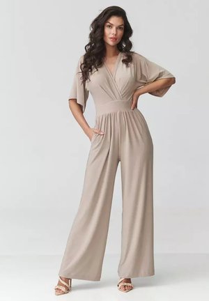 Kobieta o długich kręconych włosach, ubrana w beżowy kombinezon z szerokimi nogawkami, dekoltem w serek i krótkimi rękawami, stojąca z jedną ręką na biodrze, a drugą w kieszeni.