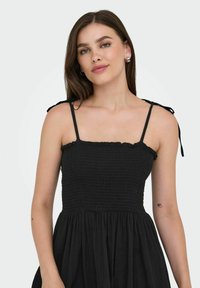 Robe noire avec des bretelles fines ajustables, un corsage smocké et une jupe évasée. Présente un décolleté à volants et un tissu texturé.