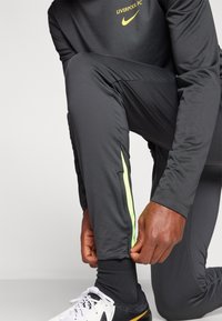 Nike Liverpool FC trainingsbroek in donkergrijs, met een contrasterende neon groene zijstreep. Materiaal is soepel en lichtgewicht. Witte schoenen.