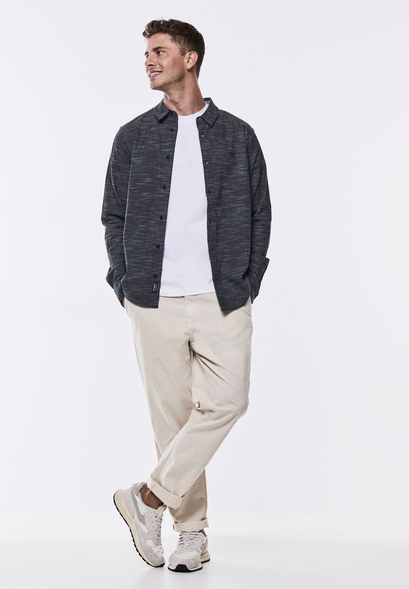 Pánská košile s dlouhým rukávem v tmavě šedé barvě, s knoflíkovým zapínáním, strukturovaný materiál, vrstvená přes bílý tričko, v kombinaci s béžovými kalhotami a šedými teniskami.