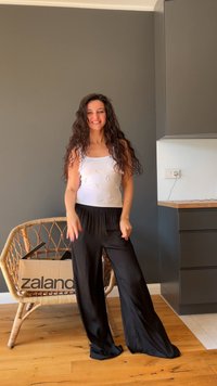 Mujer con cabello largo y rizado sonríe, vistiendo una blusa blanca sin mangas y pantalones negros sueltos, de pie en una habitación con una silla de mimbre sosteniendo una caja.