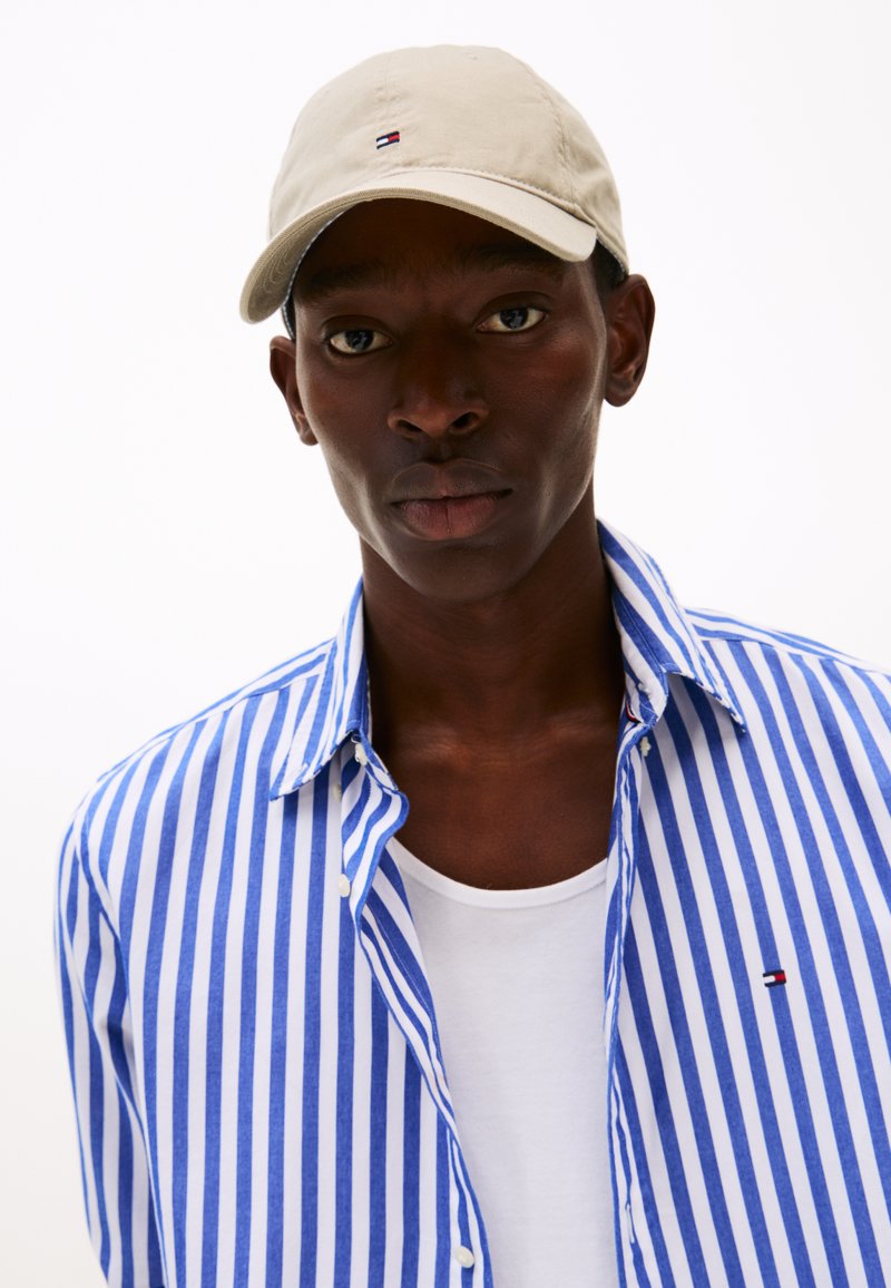 Casquette beige avec logo, chemise à manches кноптых rayures bleues et blanches, t-shirt blanc dessous. Tissu lisse, coupe ajustée, couleurs contrastées.