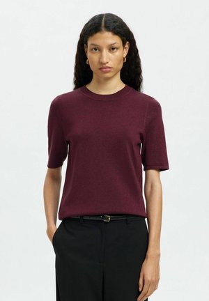 Burgunderfarbenes Kurzarm-Strickoberteil mit Rundhalsausschnitt, aus weichem Stoff gefertigt, kombiniert mit schwarzen Hosen und einem schwarzen Gürtel.