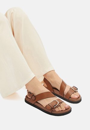 Brūnas ādas sandalēs ar plakanu zoli, ar krusteniskām siksnām un sprādzi. Tekstūrēta virsma ar atvērtu pirkstu dizainu.