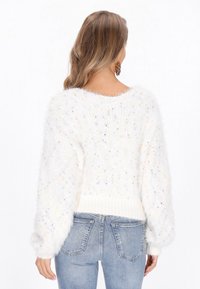 Flauschiger weißer Pullover mit mehrfarbigen Sprenkeln, breitem runden Ausschnitt und gerippten Bündchen. Getragen mit hellblauen Jeans, von hinten betrachtet.