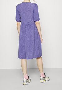 Robe violette à pois blancs. Manches courtes bouffantes, corsage ajusté et jupe évasée. Associée à des baskets grises et des chaussettes roses.