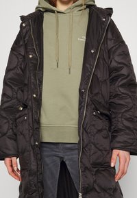 Abrigo largo acolchado negro con capucha, presenta botones a presión y una cremallera. Llevado sobre una sudadera verde con cordones. Manos del modelo visibles.