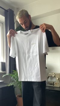 Moški s kratkimi blond lasmi in brado, ki drži navaden beli majico v notranjosti blizu okna in šivalnega stroja.