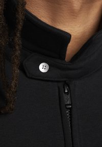 Svart zip-up kragplagg med en slät tygstruktur, med en metallknappdetalj och ett litet märkt dragband.