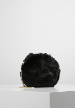 Petit sac à main rond noir en fausse fourrure avec une bandoulière chaîne dorée sur un socle blanc sur fond neutre.
