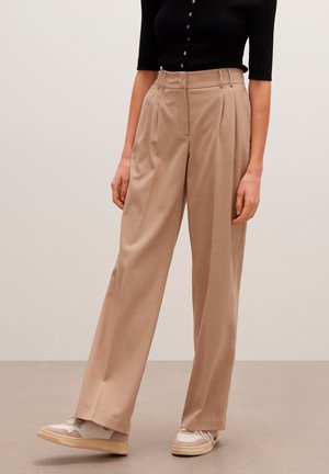 Femme porte un pantalon beige taille haute à jambes larges avec des plis, associé à des baskets beiges et blanches et un haut noir boutonné.