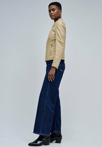 Femme se tenant de profil, portant une veste beige zippée, un jean noir à jambes larges et des bottes à talons noires, devant un fond gris uni.