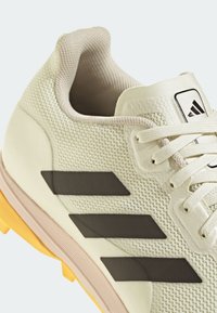 Vit sportssko med mesh-övre, beige accenter, tre svarta ränder och strukturerad sula. Har rund snörning och vadderad krage.