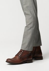 Brązowe skórzane botki sięgające do kostki z okrągłym czubkiem, sznurowane z przodu, z fakturowanymi panelami i niskim, tłoczonym obcasem, w zestawieniu z szarymi spodniami w kratę.