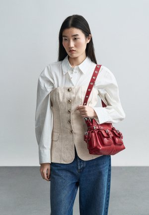 Femme aux longs cheveux foncés portant une chemise blanche à manches bouffantes et un corset beige boutonné, jean bleu, tenant un sac à main en cuir rouge sur l'épaule.