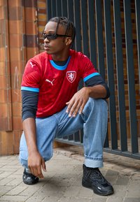 Rød sportstrøje med blå detaljer, der har Puma-logoet og et skjold. Bæres med lyseblå jeans og sorte sneakers.