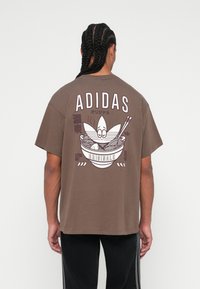 Hnědé bavlněné tričko s velkým grafickým potiskem misky nudlí s hůlkami a textem "ADIDAS" na zádech. Krátké rukávy, volný střih.