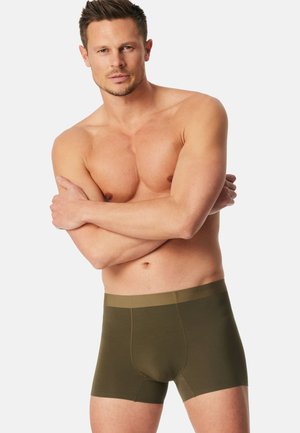 Uomo muscoloso con capelli corti che indossa boxer verde oliva, in piedi con le braccia incrociate sul petto nudo contro uno sfondo semplice.