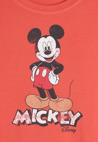 Camiseta color coral con un estampado gráfico de Mickey Mouse en blanco y negro con acentos rojos. El texto dice "MICKEY" en una fuente negrita y desgastada.