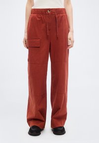 Femme portant un pantalon cargo ample de couleur rouille avec une taille bouton et cordon de serrage, associée à des baskets noires et un haut blanc.