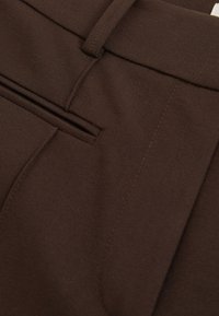 Nahaufnahme von dunkelbraunen, maßgeschneiderten Hosen mit Bund, Gürtelschlaufe, Seitentasche und Details an der Frontnaht.