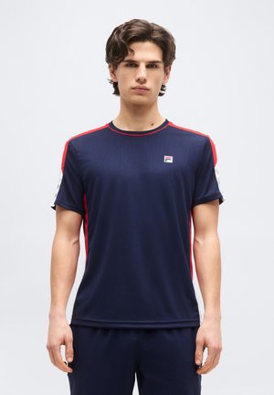 Fiatal férfi rövid, göndör hajjal, tengerészkék sportpólót visel, amelyen piros és fehér csíkok, valamint egy kis logó található a mellkason.