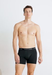 Schwarze Boxershorts mit einem elastischen Bund in Markenoptik, die eine enge Passform und eine glatte Stoffoberfläche bieten. Das Modell steht vor einem hellen Hintergrund.