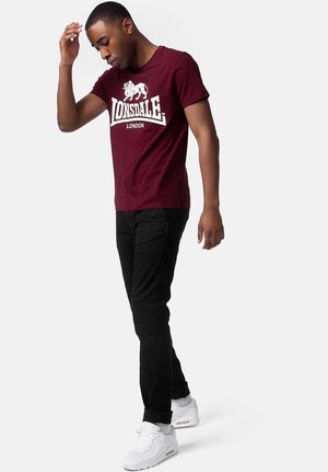 Burgundska pamučna majica s bijelim logom "Lonsdale London", uparena s crnim hlačama i bijelim tenisicama, stoji na jednostavnoj pozadini.