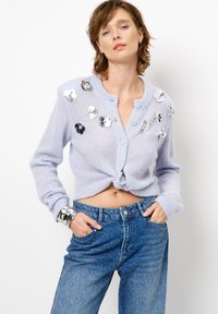 Lichtpaarse gebreide cardigan met een geknoopte voorkant, versierd met zilveren en zwarte bloemsequins, gecombineerd met een hoogwaist, blauwe spijkerbroek.