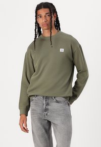 Sweatshirt vert olive avec un col rond, des manches longues et un petit patch logo blanc. Porté avec un jean gris clair présentant une texture délavée.