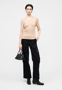 Pull beige clair en maille à col en V associé à un pantalon cargo ample noir, avec des poches et des fermetures éclair. Modèle tenant un petit sac à main noir.