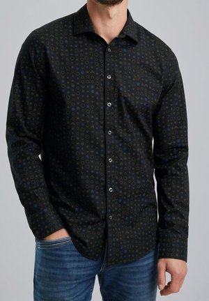 Homme portant une chemise noire boutonnée avec de petits motifs circulaires et triangulaires en bleu foncé et gris, associée à un jean bleu.